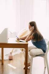 A young attractive student talks on a smartphone while studying at home, discusses homework or a joint project with friends, makes notes in a notebook