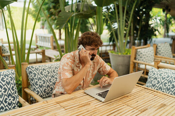 Handsome man with beard using lap top and mobyle phone , sitting in outdoor  cafe with tropical interior.