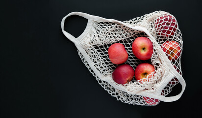 Top view of a black background on which lies a white string bag with big ripe juicy red apples, flat lay copy space.
