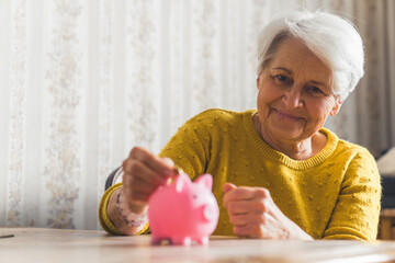 Optimistic happy smiling caucasian senior woman putting coins into a piggy bank. Savings. High...