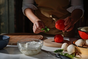 Close-up Hands of housewife, chef holding ripe tomato and basil leaves while preparing delicious meal in rustic kitchen