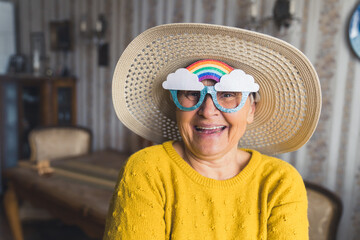 Funny caucasian old single woman wearing paper party cartoon glasses with rainbow and clouds on...