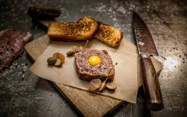 Decorated dish with toasts and tartare. Raw egg on top of meat. Knife and meat on a black stone table. Salt and pickles on table. 