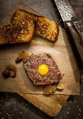Decorated dish with toasts and tartare. Raw egg on top of meat. Knife and meat on a black stone table. Salt and pickles on table. 