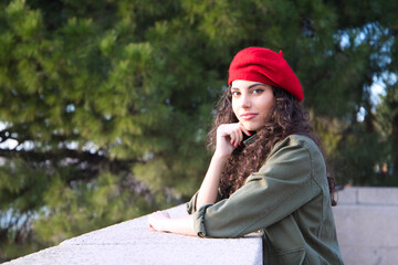 Beautiful young latina woman with curly brown hair wearing a red cap and dressed in casual clothes is sightseeing in Europe. She is posing for photos. Mediterranean pine tree in the background.Tourism
