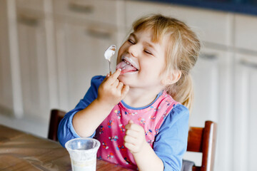Little toddler girl eating yogurt for breakfast. Cute healthy baby sitting in the kitchen or at nursery and having meal.