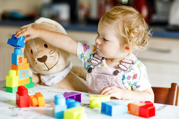 Adorable toddler girl with favorite plush bunny playing with educational toys in nursery. Happy healthy child having fun with colorful different plastic blocks at home. Cute baby learning creating.