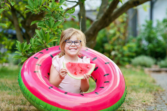 Cute little girl with glasses eating watermelon on inflatable ring in summertime. Happy smiling preschool child having fun. Healthy summer food and snacks for kids.