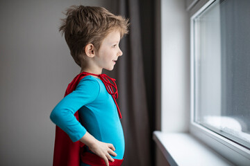 Stay at home. Little superhero boy in a red cape alone looks sadly out the window
