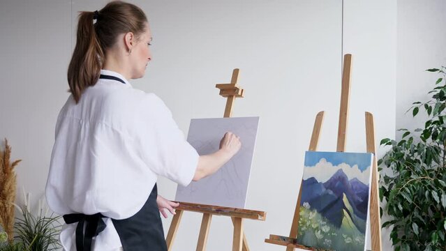 Mature woman artist redraws mountains on easel canvas against white wall. Skilled lady recreates picture of landscape using pencil closeup