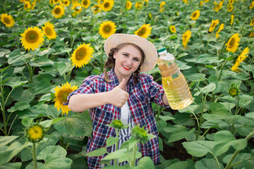 Beautiful smiling joyful middle-aged farmer woman holding bottles of golden sunflower oil in her hands and  thumbs up gesture in a harvest field of sunflowers on a sunny day