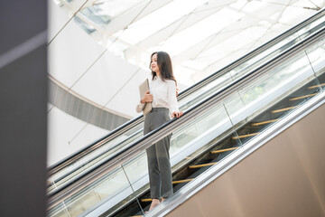 Young business woman holding a briefcase in hand riding an escalator - obrazy, fototapety, plakaty