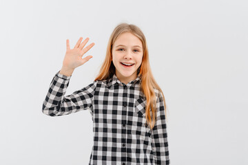 Friendly teen girl says hello, waves hand and smiles happy at you, stands over white background in...