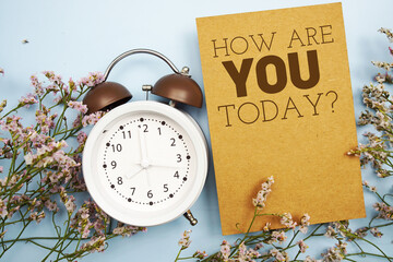 How are you today? written on paper card with flower and alarm clock on blue background