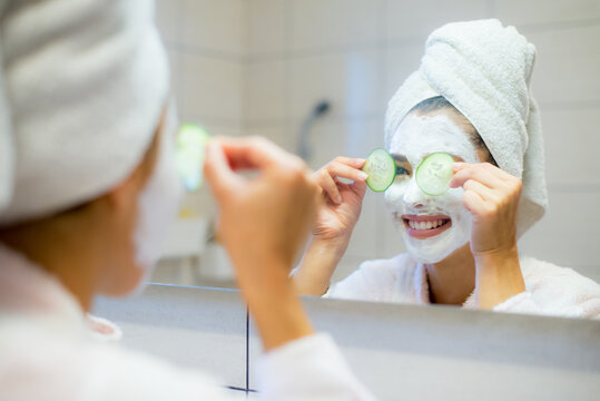 Young beautiful woman in bathrobe putting facial mask on face in front of the bathroom mirror