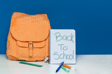 orange backpack with front pocket on blue background with markers, ruler and pen on white table....