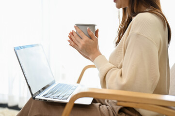 Relaxed Asian female sits on armchair in the living room, holding a coffee mug and using laptop.