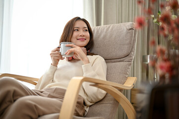 Relaxed and chill young Asian woman is laying on her armchair having a morning coffee