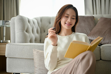 Attractive young Asian woman sits in the minimal living room enjoying reading a book.