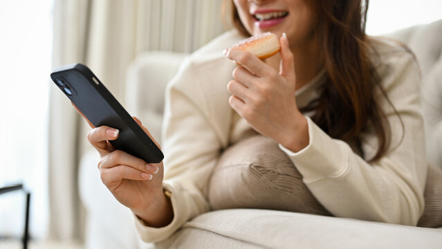 Asian female laying on comfy sofa, eating doughnut and using smartphone. cropped image