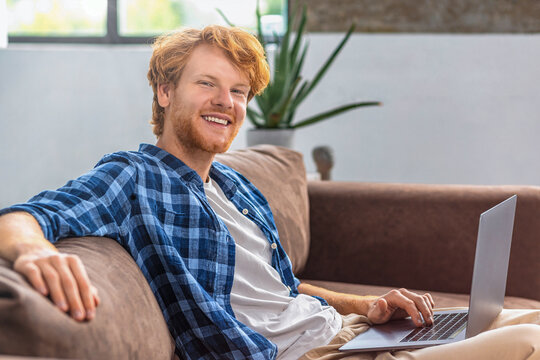 Successful businessman or freelancer working using computer laptop sitting sitting on sofa in modern office. Young bearded redhaired  man entrepreneur looking at the camera and smiling