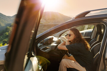 Happy young woman driver traveler in sunglasses sitting in car enjoying sunset while traveling