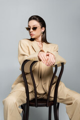 young woman in stylish beige outfit and sunglasses sitting on wooden chair isolated on grey