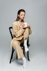 full length of brunette woman in stylish beige suit holding cup of coffee and sitting on wooden chair on grey