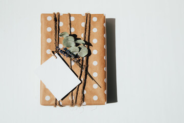 The gift is wrapped in kraft paper with white circles, tied with twine, decorated with dry flowers with postcard. Top view