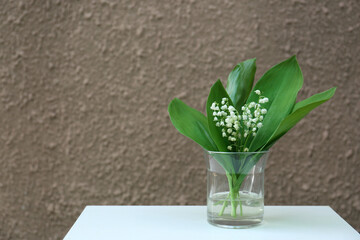 Beautiful lily of the valley flowers in glass vase on white table near brown wall, space for text