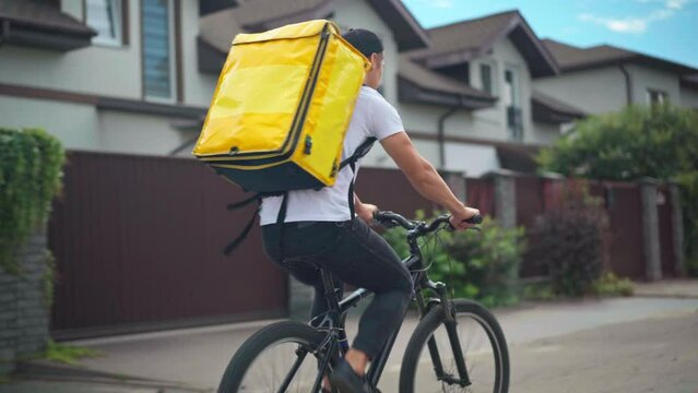 Tracking shot of young Caucasian man with yellow delivery bag riding bicycle on suburban street. Live camera follows delivery boy delivering food and drink on summer day outdoors. Service concept