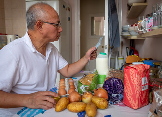 A senior man in the kitchen checking the cost of daily essential food as rising inflation and cost...