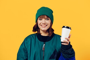 a happy, joyful woman stands on a yellow background in a stylish jacket and holding a coffee glass in her hands, smiling pleasantly while looking at the camera