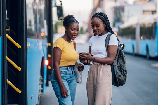 Two african american woman waiting for a bus and using a smartphone