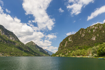 Green waters of the lake (Lago d'Idro) framed by alpine rocks with evergreen trees under a clear blue sky with white clouds, wide panorama. Brescia, Lombardy, Italy