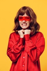 a funny, charming woman stands on a yellow background in a stylish red shirt and fashionable glasses, holding her hands in a fist near her chin and happily winking at the camera