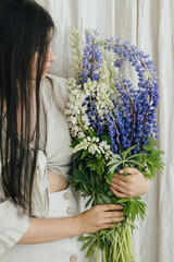 Young female in linen dress posing with lupine flowers. Gathering and arranging summer wildflowers at home in countryside. Stylish woman holding lupine bouquet in rustic room, close up