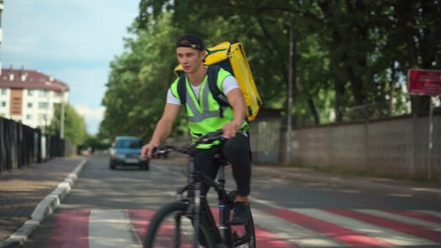 Concentrated delivery boy searching address riding bike in residential neighborhood gesturing thumb up giving way turning the road. Front view dolly shot Caucasian young man delivering order
