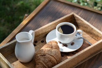 A cup of black coffee, milk and a croissant on a wooden table against the background of nature.