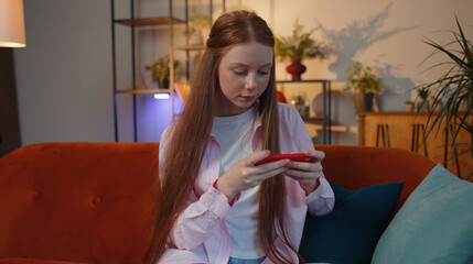 Worried young redhead teen child girl enthusiastically playing racing shooter online video games on mobile phone. Red hair kid using smartphone gadget app with drive simulator at home sitting on couch