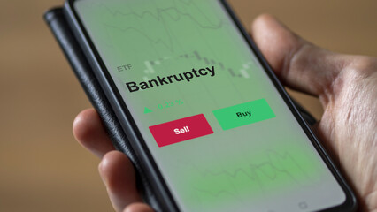 An investor's analyzing the bankruptcy etf fund on a screen. A phone shows the prices of Bankruptcy