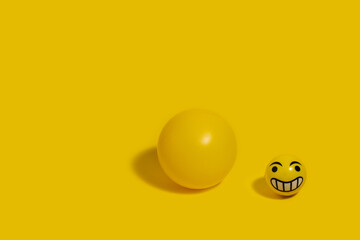 Small yellow bouncy ball with a smiley emoticon next to a big yellow ball on a yellow background.