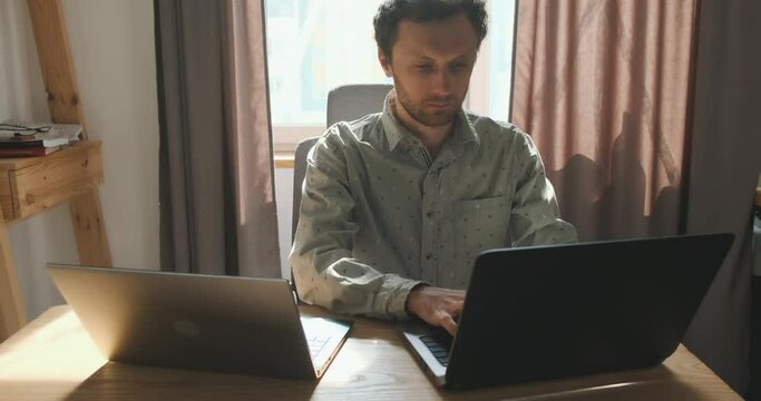 Young IT specialist works on two laptops at the same time. Front view, portrait, home office, desktop. The concept of the modern world, online remote work, how to do everything, overstrain.