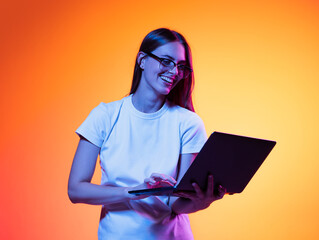 Portrait of young smiling girl, student in white t-shirt with laptop isolated on orange color background in neon light. Concept of beauty, fashion, emotions