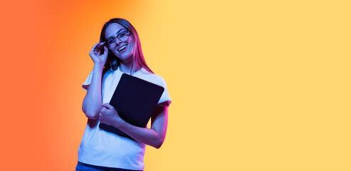 Portrait of young smiling girl, student in white t-shirt with laptop isolated on orange color background in neon light. Concept of beauty, fashion, emotions