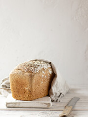 Loaf of rye and wheat sourdough bread covered by beige linen kitchen towel on a white cutting board. Bread knife on a wooden table. Space for text