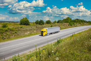 yellow cargo truck on the highway. asphalt road among green fields and beautiful clouds. cargo delivery and transportation concept