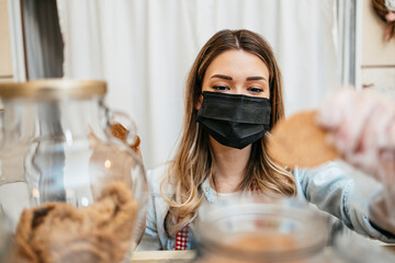 Beautiful young female seller with protective face mask working in a confectionery shop or bakery....