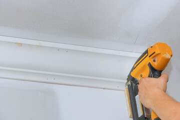 Carpenter installing ceiling corner crown molding with the help of an air nail gun to new house