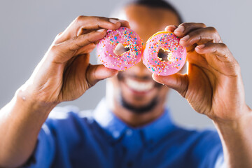 Smiling african american man holding fresh pink donuts in front of face against gray background
